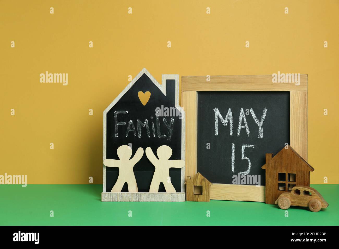 Alles Gute Zum Internationalen Familientag. Kleine Tische mit Text, Hausmodellen, Personenfiguren und Spielzeugauto auf einem grünen Tisch vor gelbem Hintergrund Stockfoto
