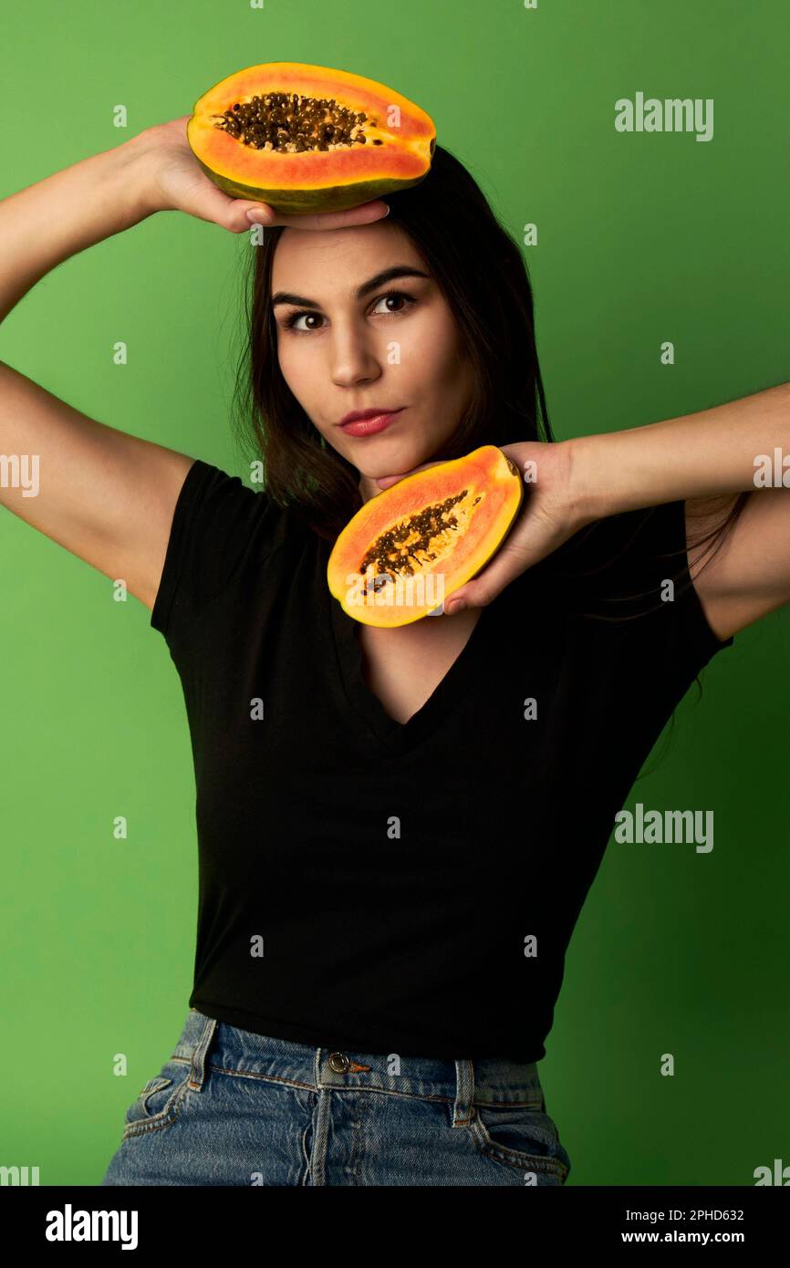 Eine Frau, die eine geschnittene Papaya in einem schwarzen Hemd hält, vor einem grünen Hintergrund steht und in die Kamera schaut Stockfoto
