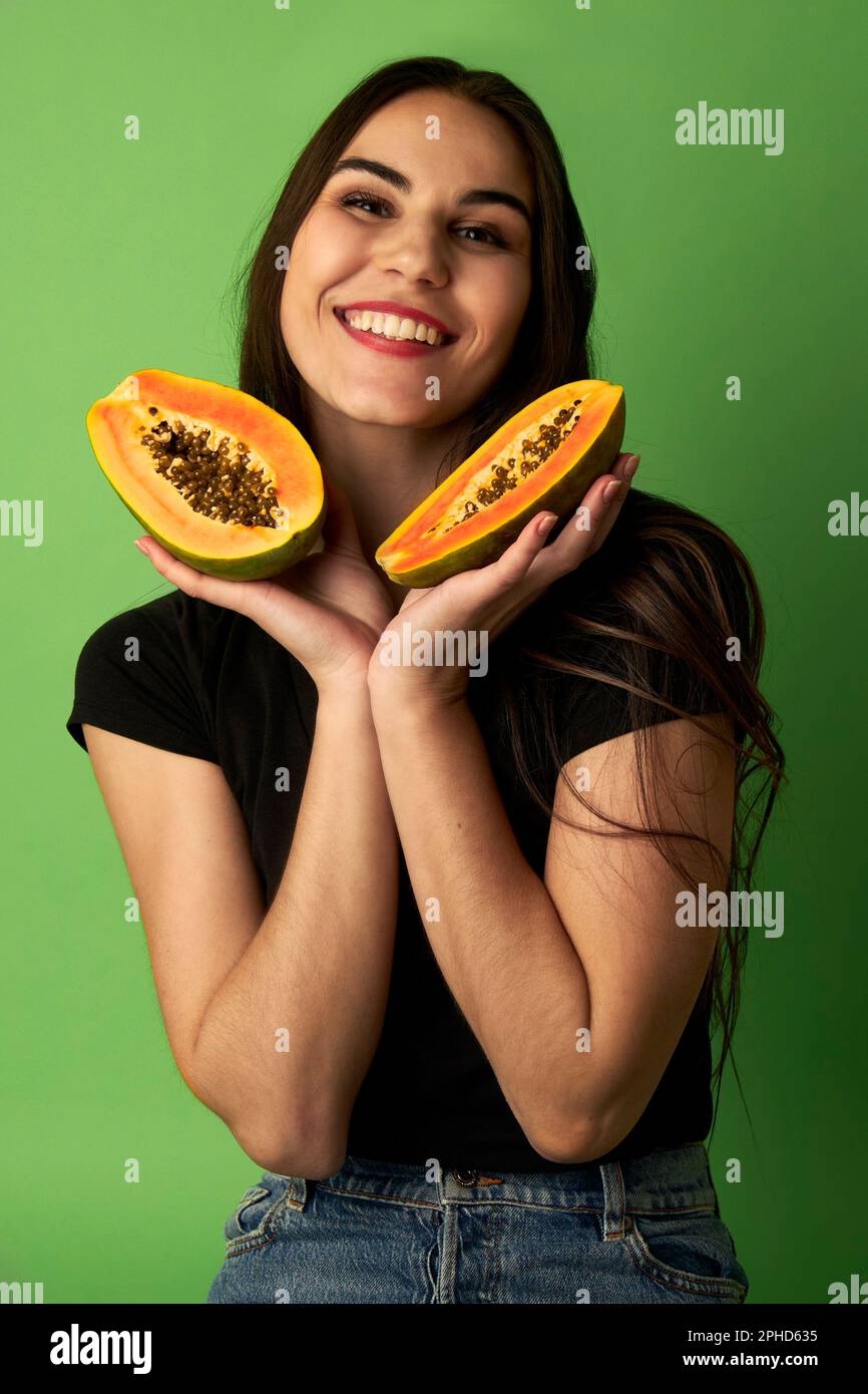 Eine Frau, die eine geschnittene Papaya in einem schwarzen Hemd hält, vor einem grünen Hintergrund steht und vor der Kamera lächelt Stockfoto