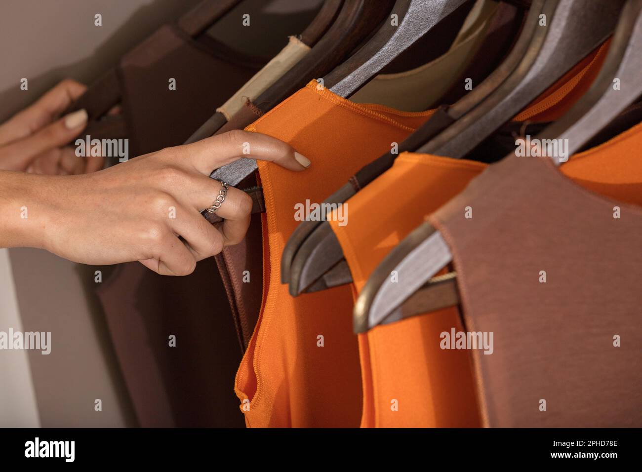 Nahaufnahme der Hand einer gut gepflegten Frau, die Kleidung in einem Kleiderschrank oder Laden aussucht, mit ordentlich aufgehängten Outfits an Kleiderbügeln in Orange-, Braun- und B-Farbtönen Stockfoto