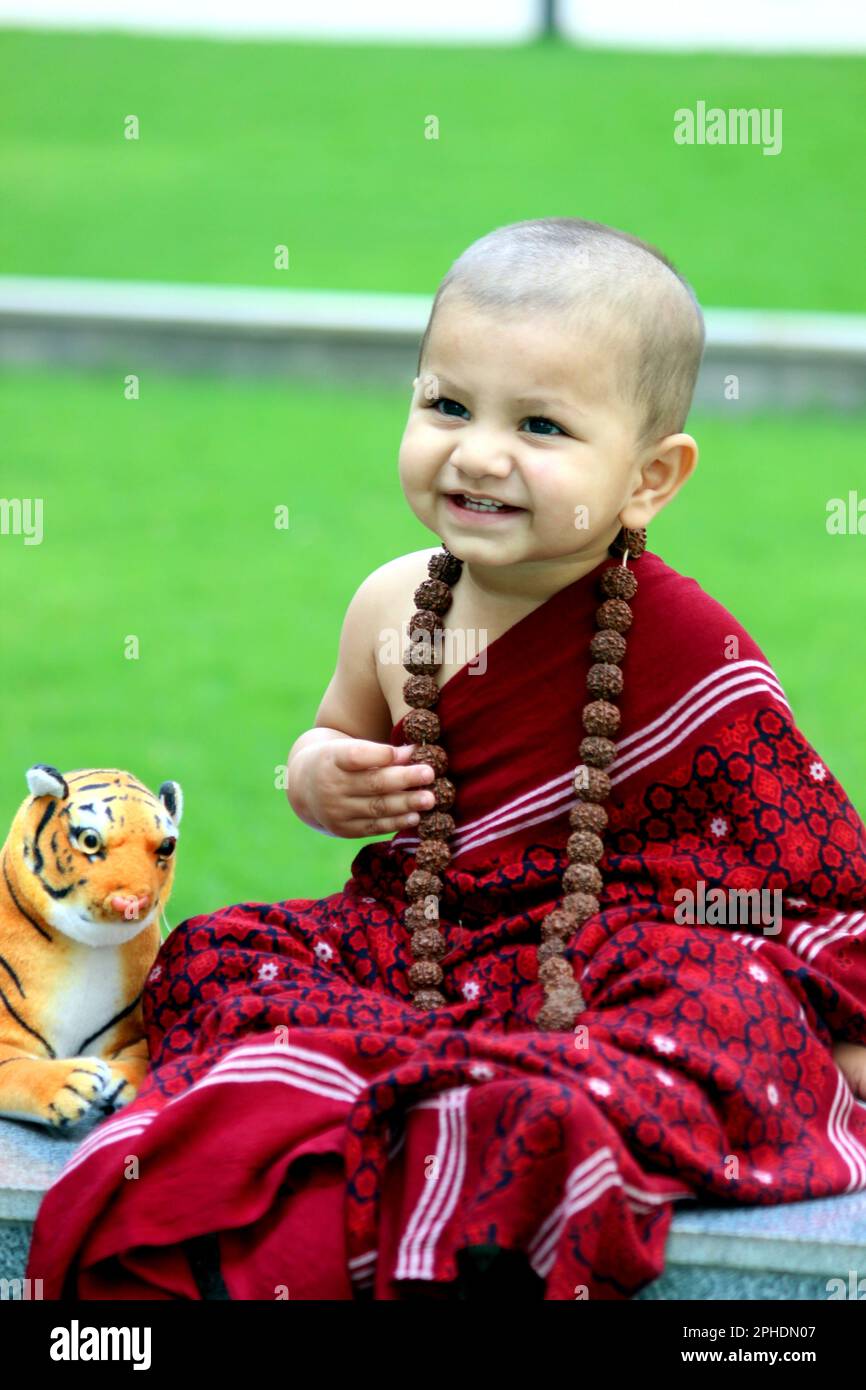 Ein süßer, kahlköpfiger Junge trägt Monk Avatar, einen kastanienbraunen Schal, Rudraksh, und ein süßes Lächeln. Ein Tiger-Spielzeug ist neben dem Jungen. Stockfoto