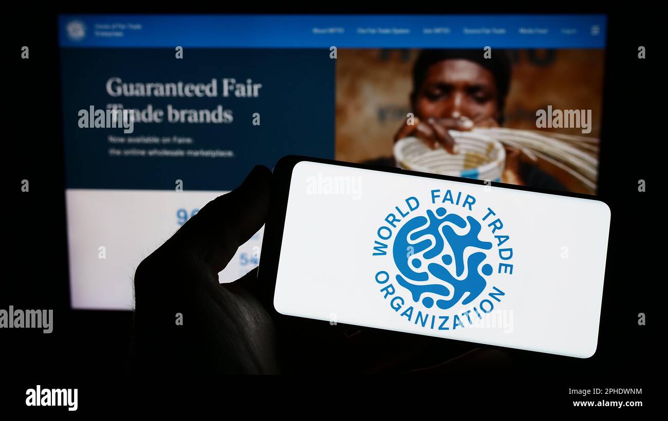 Person, die ein Mobiltelefon mit dem Logo der Welthandelsorganisation (WFTO) auf dem Bildschirm vor der Webseite hält. Konzentrieren Sie sich auf das Display des Telefons. Stockfoto