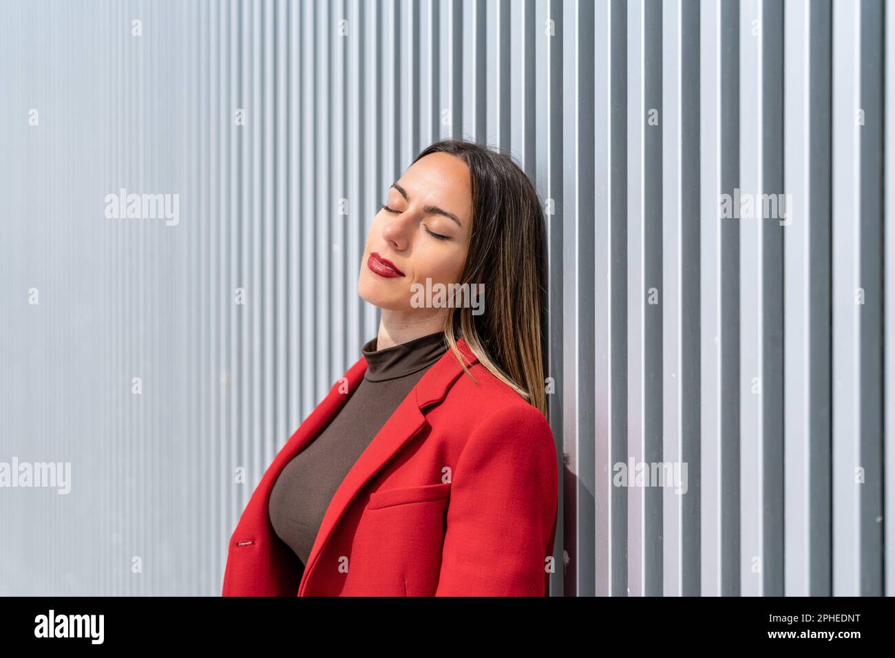 Junge Frau in rotem Mantel und dunkelblondem Haar, die sich an einer grauen Wand lehnt, mit geschlossenen Augen an sonnigen Tagen Stockfoto