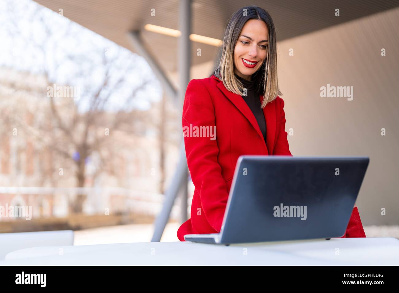 Glückliche junge Frau mit dunkelblondem Haar und rotem Lippenstift, die am Tisch sitzt und vor verschwommenem Hintergrund auf den Laptop-Bildschirm schaut Stockfoto