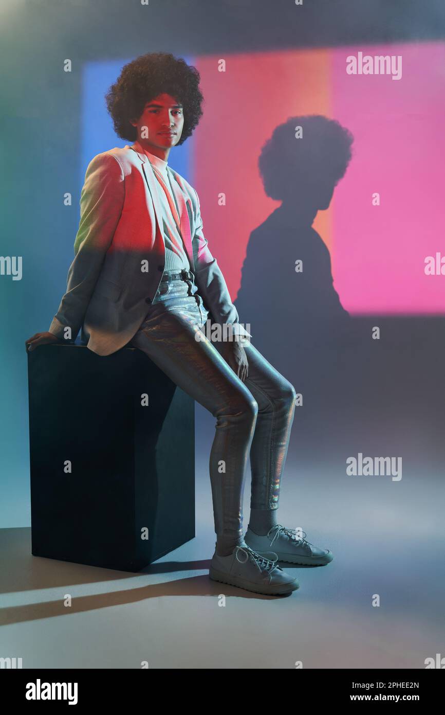 Der ganze Körper eines jungen hispanischen männlichen Models in elegantem Freizeitanzug sitzt auf einem schwarzen Würfel vor der bunten, gestreiften Beleuchtung des Projektors Stockfoto