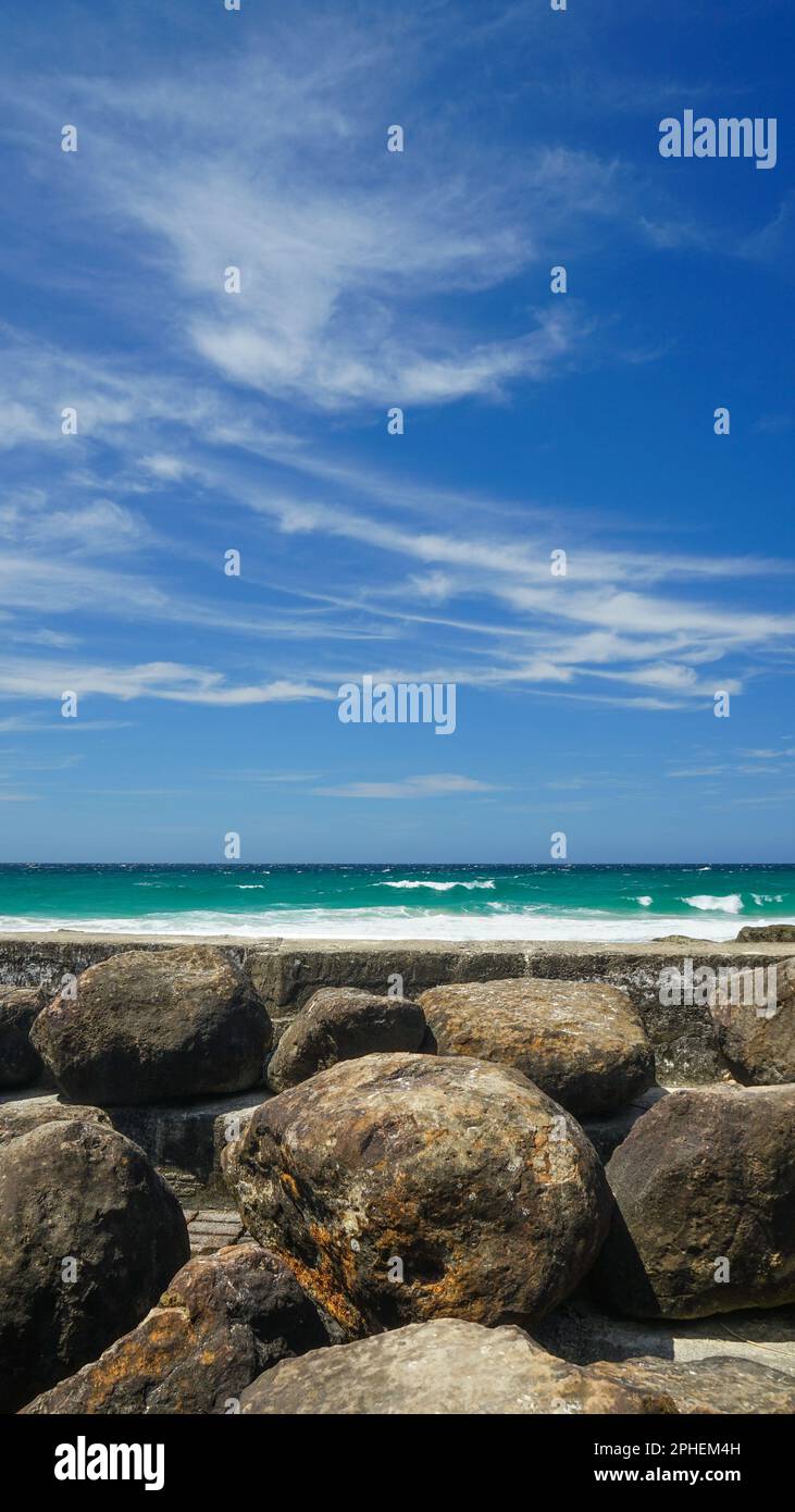 Mit Blick über eine Betonwand auf das Meer und den blauen Himmel mit weichen Wolken. Felsbrocken im Vordergrund. Snapper Rocks, Coolangatta. Stockfoto