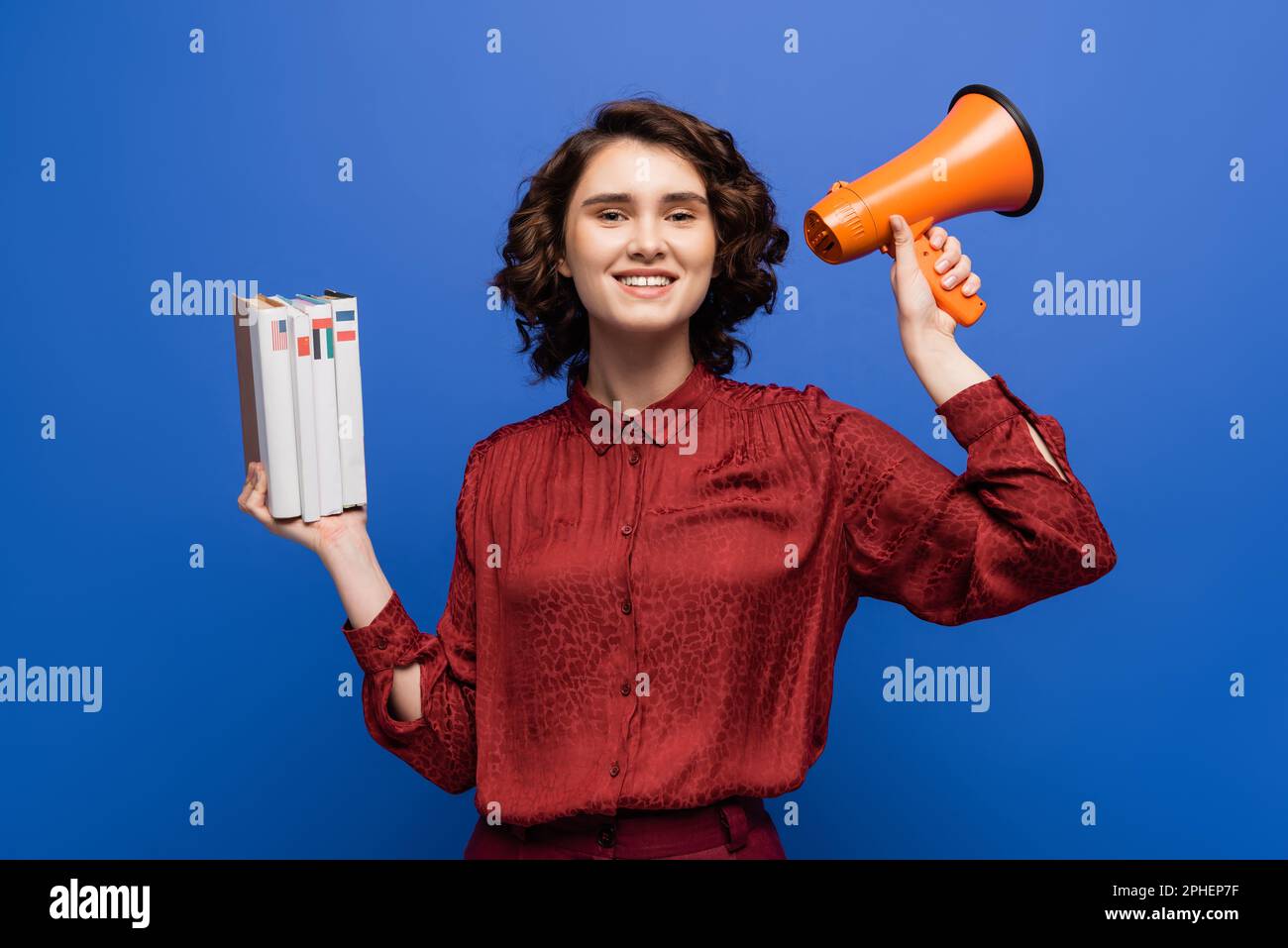 Junger und fröhlicher Sprachlehrer mit Megafon und Lehrbüchern isoliert auf blauem Stockbild Stockfoto