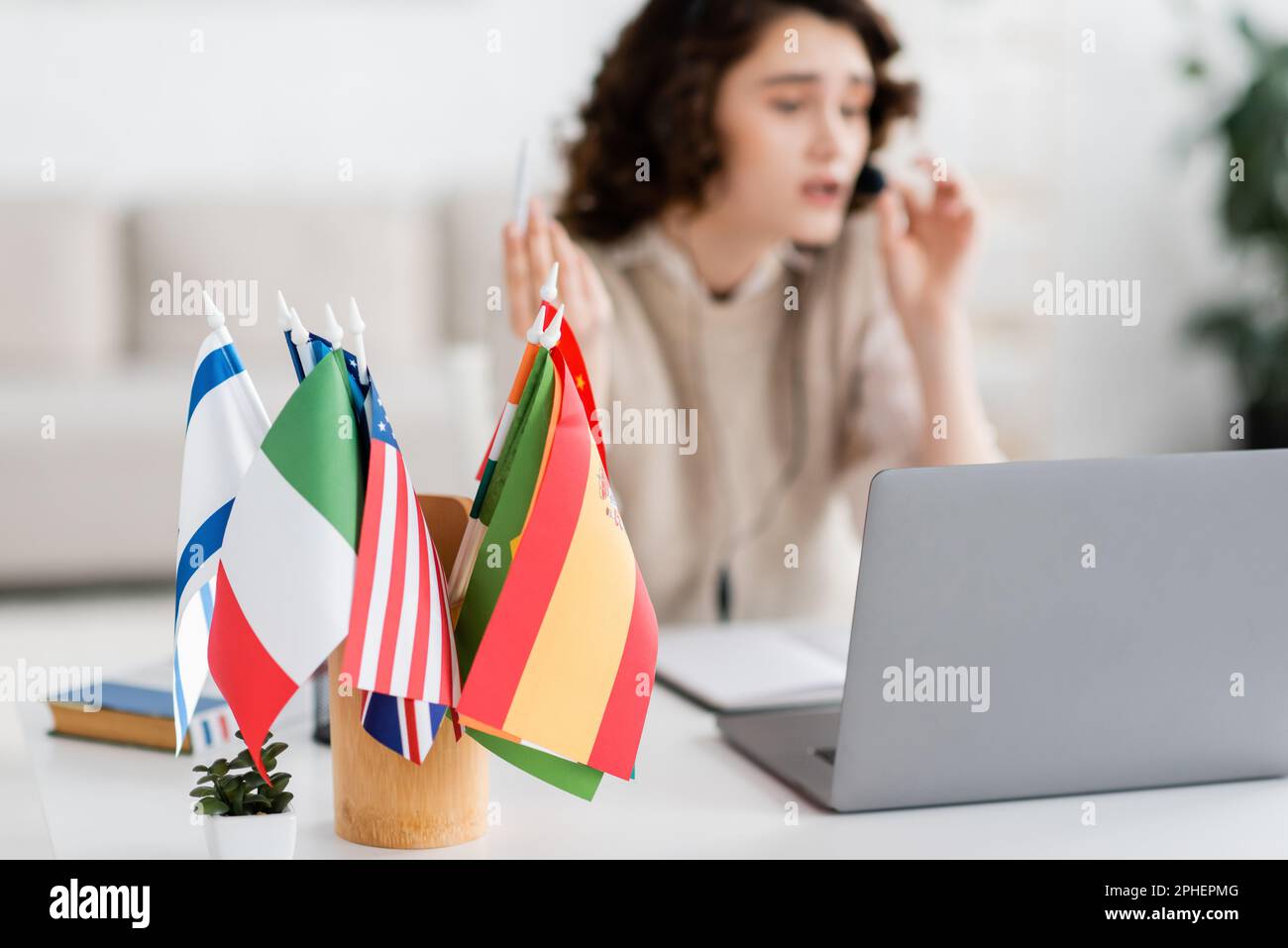 Selektiver Fokus auf verschiedene internationale Flaggen in der Nähe von Laptops und verschwommener Sprachlehrer, die zu Hause arbeiten, Stock Image Stockfoto