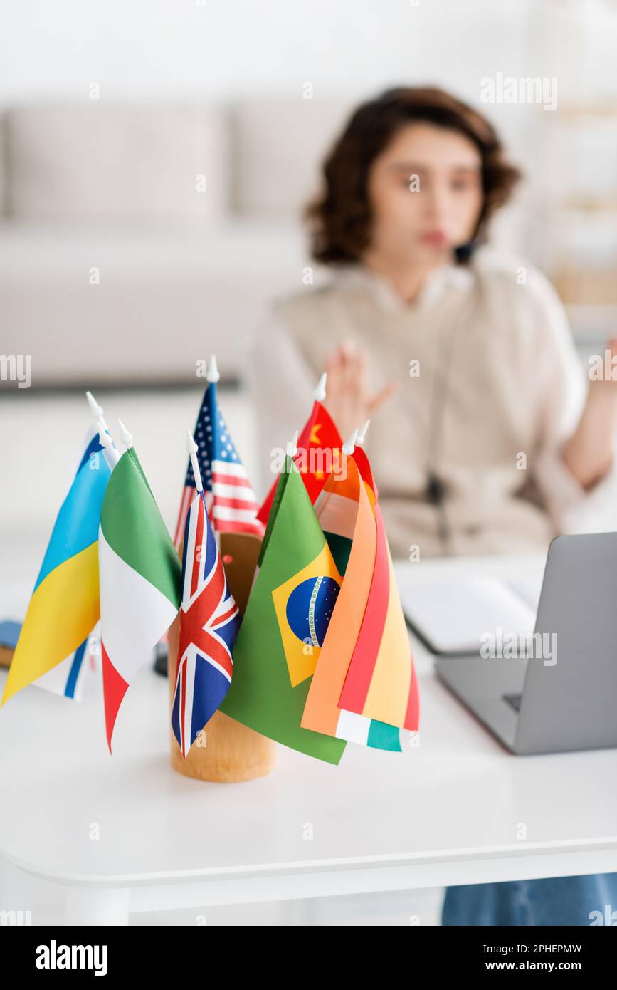 Selektiver Fokus auf internationale Flaggen in der Nähe von verschwommenem Laptop und jungen Sprachlehrern, die zu Hause arbeiten, Stock Image Stockfoto