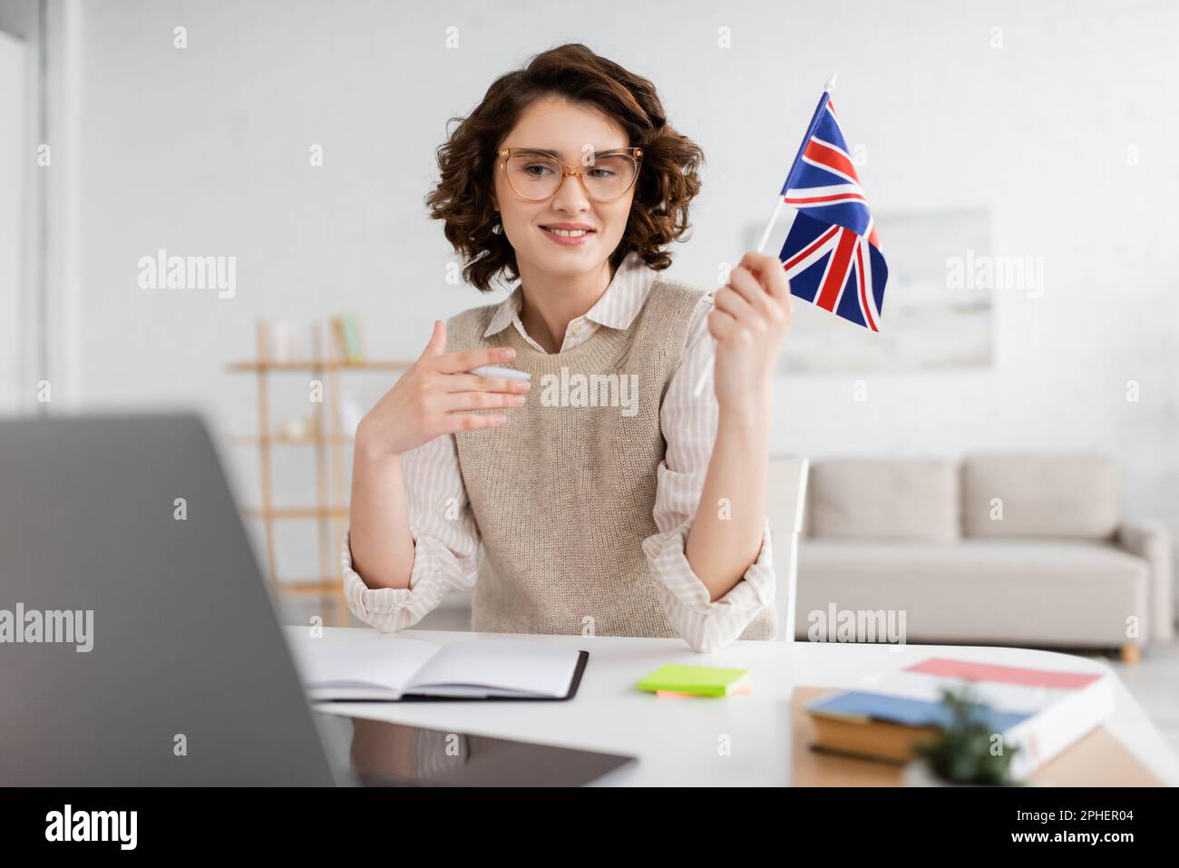 Junger Sprachlehrer mit Brille hält Flagge von Großbritannien in der Nähe des Laptops auf verschwommenem Vordergrund zu Hause, Stockbild Stockfoto