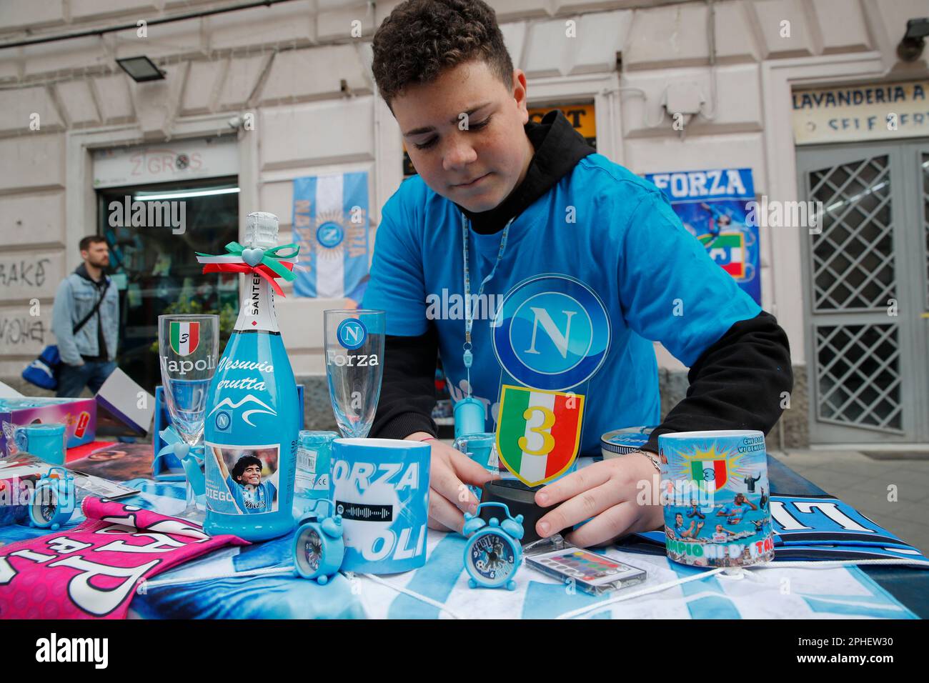 Neapel, Italien. 11. März 2023. Ein Junge stellt einen Stand mit Champagnerflaschen auf, in dem das Bild von Diego Armando Maradona, Flaggen und andere Gegenstände vor einem Geschäft ausgestellt sind. In der Stadt beginnen die Vorbereitungen, um den Sieg der Fußballliga der Serie A von SSC Napoli zu feiern, die derzeit den ersten Platz in der Rangliste belegt, mit 18 Punkten vor dem zweiten, wobei noch 12 Spiele zu spielen sind. Nach den Siegern in den Jahren 1987 und 1990 wäre dies die dritte Trophäe von Neapel in der italienischen Spitzenliga Scudetto, mit Diego Armando Maradona auf dem Spielfeld. Kredit: Unabhängige Fotoagentur/Alamy Live News Stockfoto