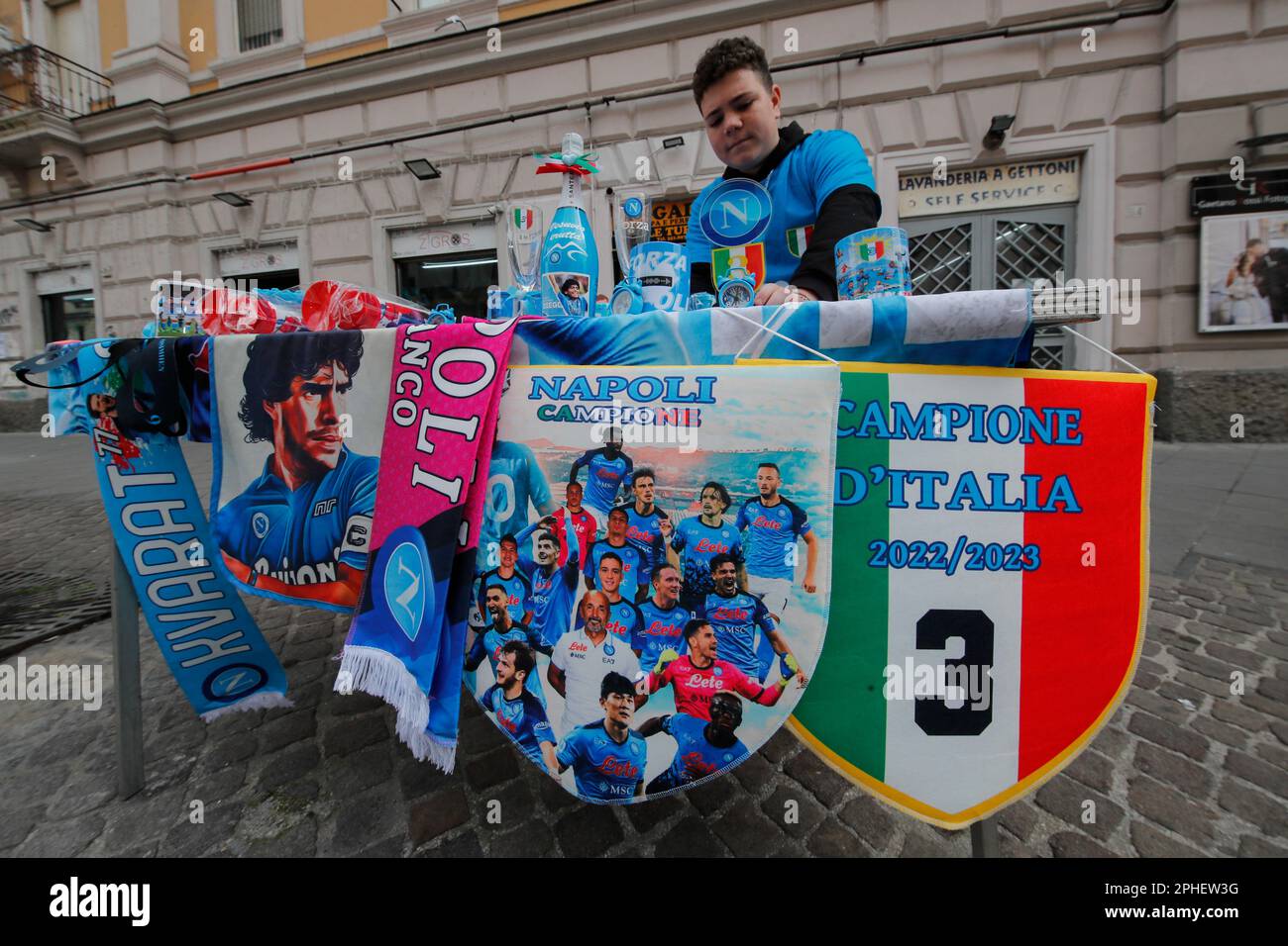 Neapel, Italien. 11. März 2023. Ein Junge stellt einen Stand mit Champagnerflaschen auf, in dem das Bild von Diego Armando Maradona, Flaggen und andere Gegenstände vor einem Geschäft ausgestellt sind. In der Stadt beginnen die Vorbereitungen, um den Sieg der Fußballliga der Serie A von SSC Napoli zu feiern, die derzeit den ersten Platz in der Rangliste belegt, mit 18 Punkten vor dem zweiten, wobei noch 12 Spiele zu spielen sind. Nach den Siegern in den Jahren 1987 und 1990 wäre dies die dritte Trophäe von Neapel in der italienischen Spitzenliga Scudetto, mit Diego Armando Maradona auf dem Spielfeld. Kredit: Unabhängige Fotoagentur/Alamy Live News Stockfoto