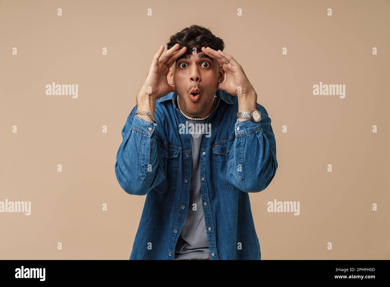 Junger, attraktiver, schockierter Mann mit offenem Mund und weit geöffneten Augen, der seinen Kopf berührt und in die Kamera schaut, während er über isoliertem Braun steht Stockfoto