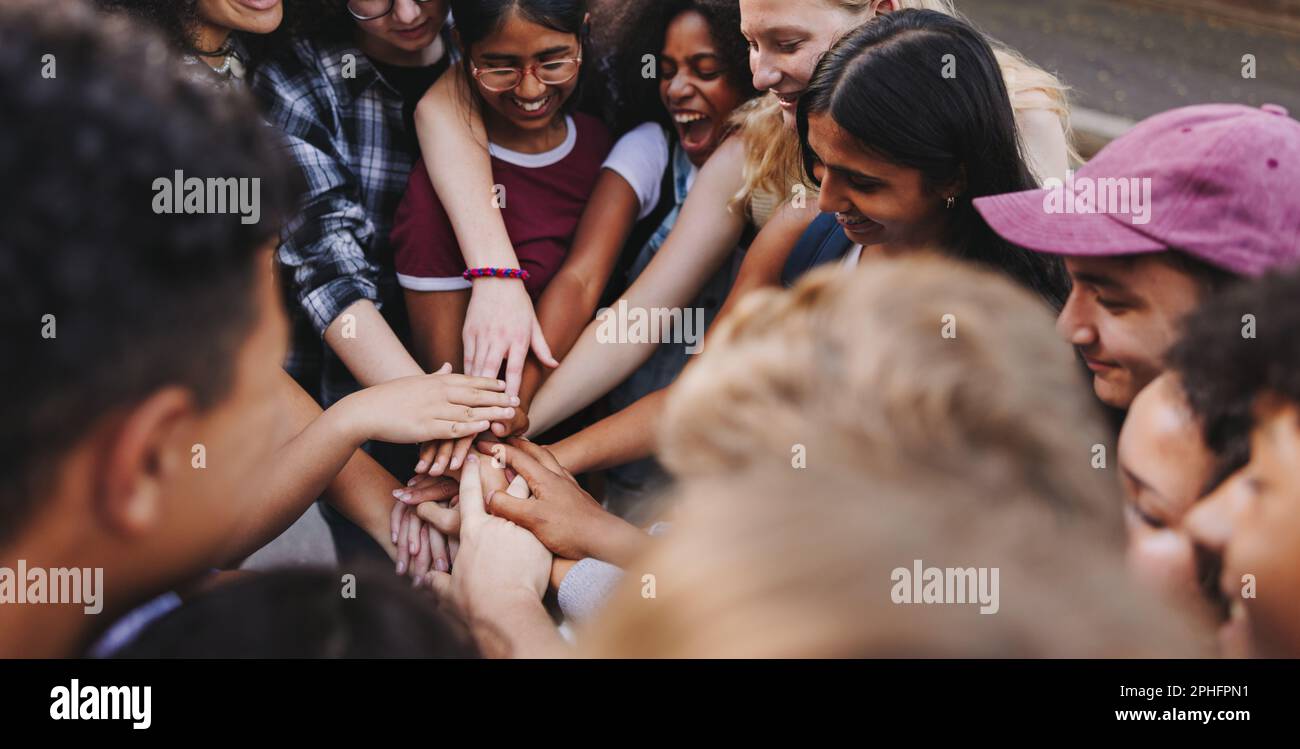 Multikulturelle Teenager, die fröhlich lächeln, während sie ihre Hände in einem Zusammentreffen zusammenlegen. Eine Gruppe unterschiedlicher junger Menschen, die Einheit und Teamarbeit symbolisieren. Stockfoto