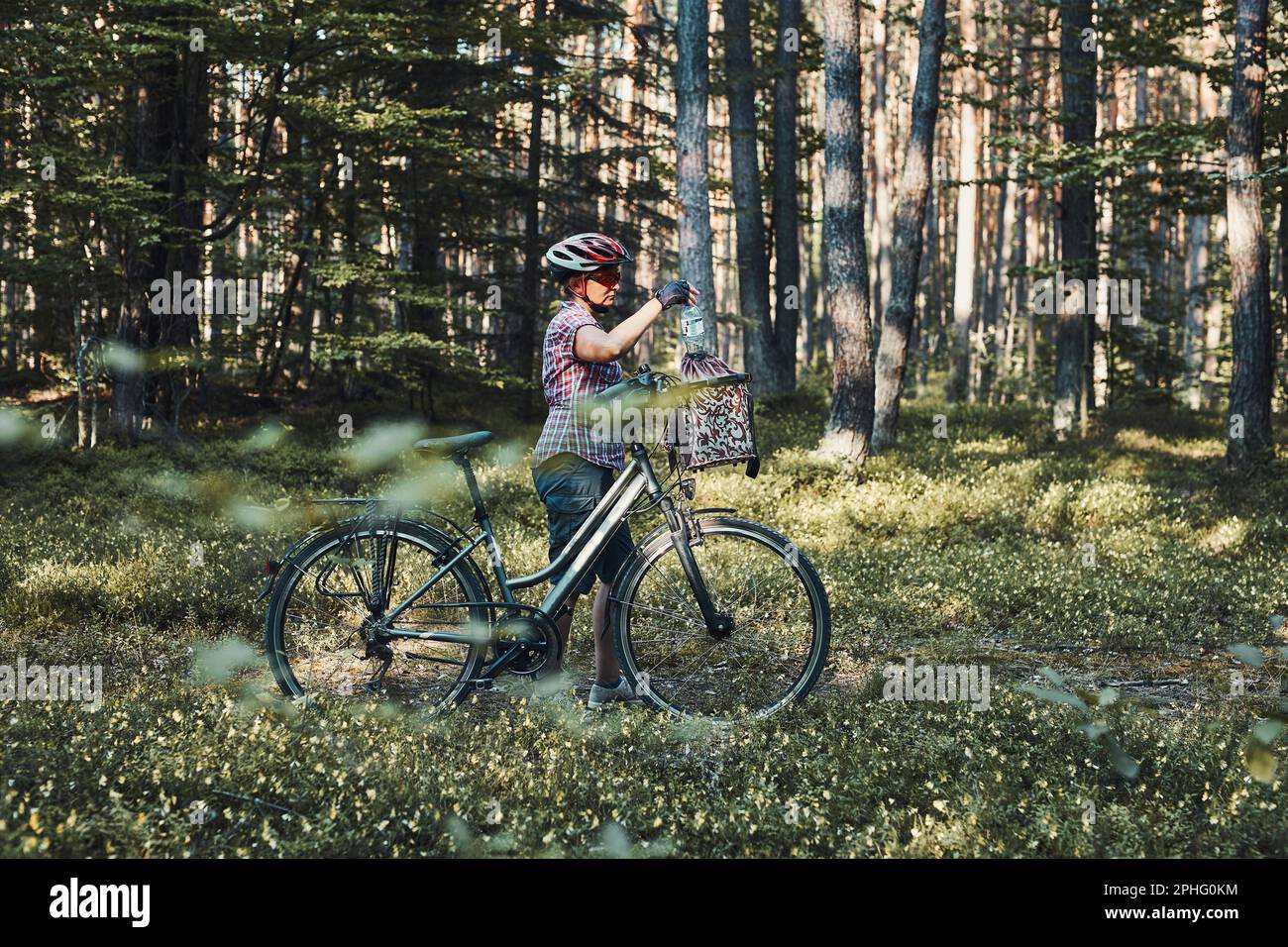 Aktive Frau verbringt freie Urlaubszeit auf einem Fahrradausflug in einem Wald an einem sonnigen Sommertag. Frau mit Fahrradhelm und Handschuhen, die das Fahrrad halten Stockfoto