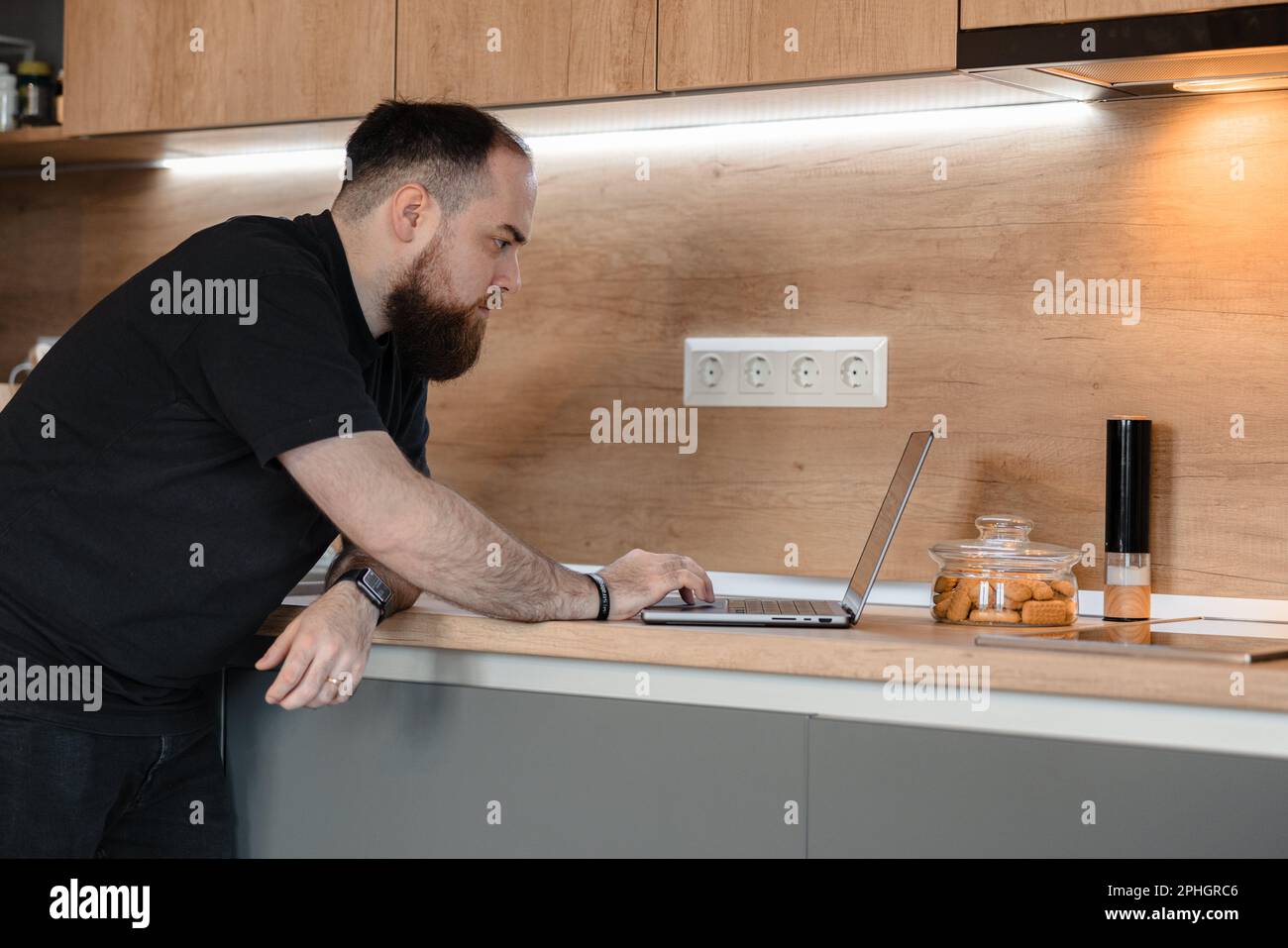 Bärtiger Mann, der bequem in seiner eigenen Küche an seinem Laptop arbeitet Stockfoto