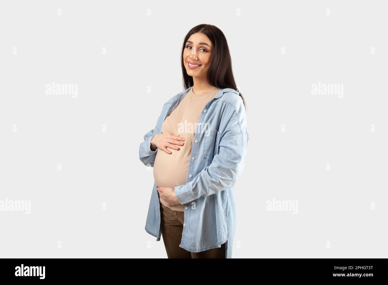Süße schwangere Dame, die auf ihr Baby wartet, in Freizeitkleidung posiert, vor die Kamera schaut und lächelt, weißer Hintergrund, freier Platz Stockfoto