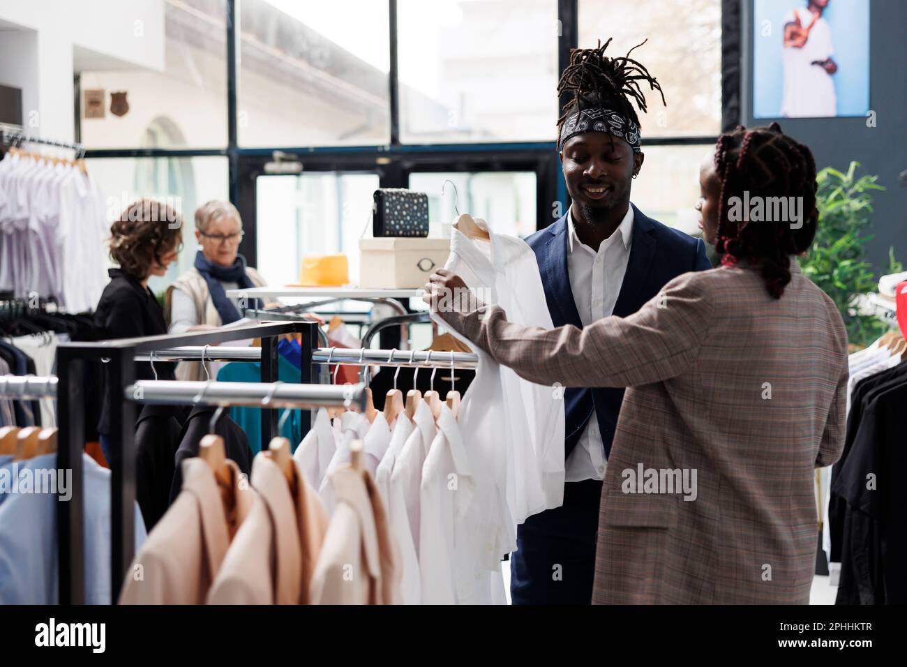 Mitarbeiter im Ausstellungsraum hilft afroamerikanischen Kunden bei der Auswahl des weißen Hemds während der kommerziellen Aktivitäten. Eleganter Kunde, der modische Kleidung im Einkaufszentrum kauft. Modekonzept Stockfoto