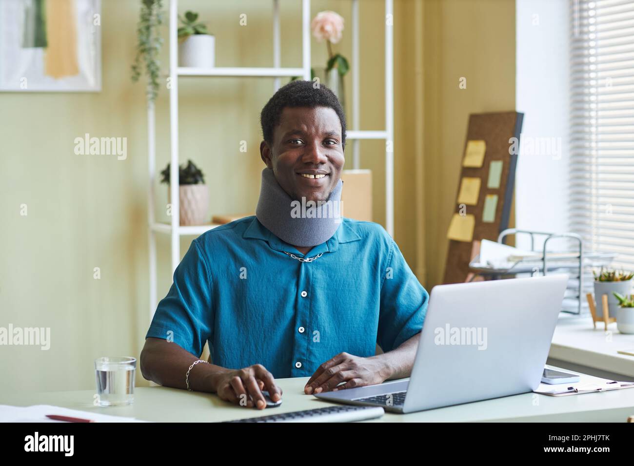 Porträt eines schwarzen jungen Mannes mit Nackenstütze, der am Schreibtisch im Büro arbeitet und vor der Kamera lächelt Stockfoto