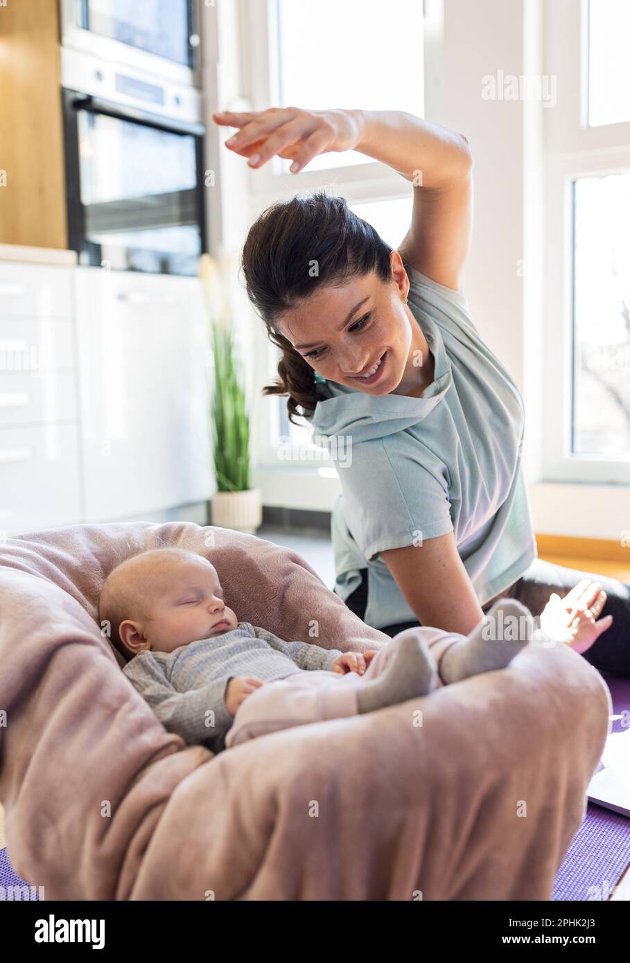 Junge, hübsche Mutter, die zu Hause vor Küchenschränken Yogamatte benutzt und ihr Neugeborenes beim Schlafen beobachtet. Geistig und körperlich gesund Stockfoto