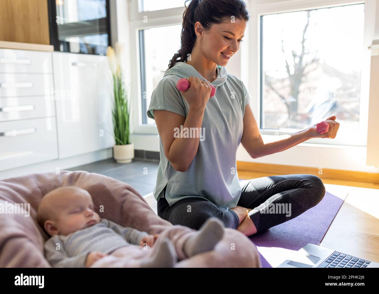 Die junge, hübsche Mutter schaut auf den Laptop und trainiert zu Hause mit Kurzhanteln auf der Yoga-Matte, während ihr neugeborenes Baby neben ihr schläft. Gesundes geistiges und Stockfoto