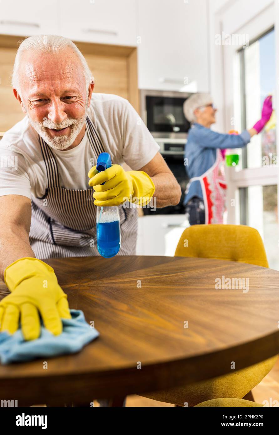 Ein reifes Paar, das zu Hause Hausarbeiten macht, ein Mann, der den Esstisch abwischt, während eine Frau die Fenster wäscht Stockfoto