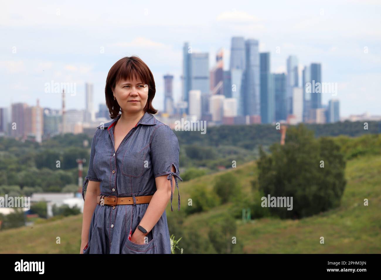 Porträt einer Frau in Jeanskleid, die auf einem Hügel im Hintergrund von Moskau steht. Sommerreisen und Freizeit, urbane Landschaft mit Wolkenkratzern Stockfoto