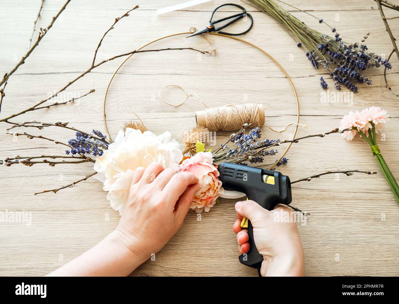 Frauenhände machen einen modernen Kranz im Frühlingsdesign mit Blumenmotiv zur Innendekoration. Runder Metallbügelrahmen mit verschiedenen realen und künstlichen Zweigen Stockfoto