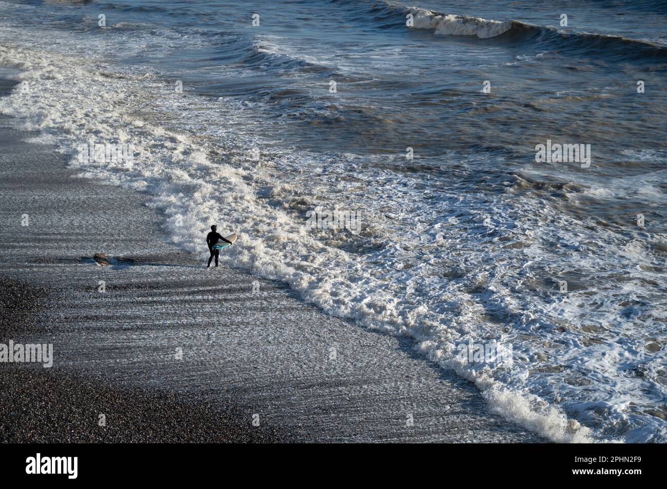 Ein Mann mit einem Surfbrett, der im Birling Gap, West Sussex UK, auf Wintersurfen wartet Stockfoto