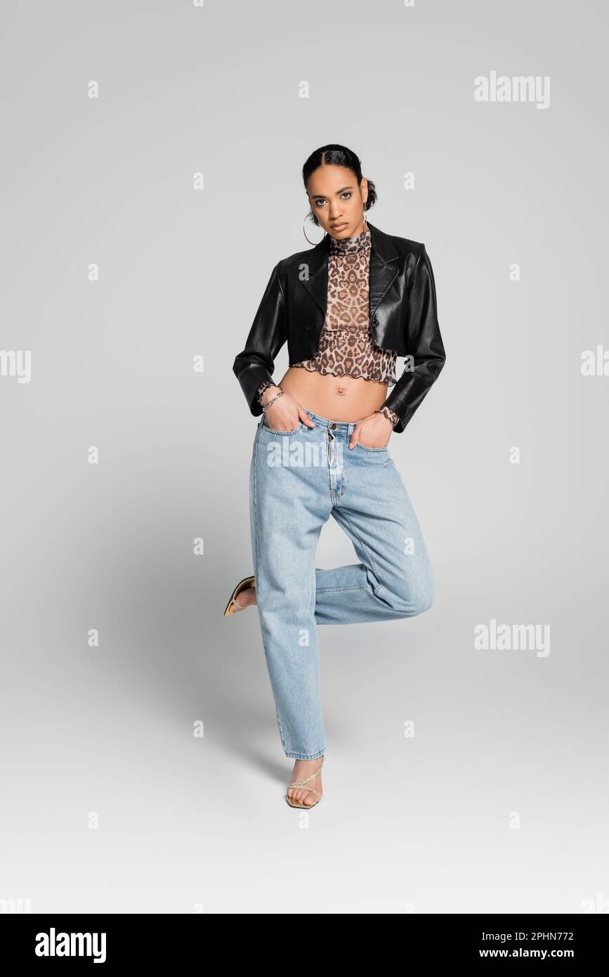 Die gesamte Länge des afroamerikanischen Modells in gekürzter Jacke posiert mit Händen in Taschen, während man auf einem Bein auf einem grauen, Stock Bild steht Stockfoto