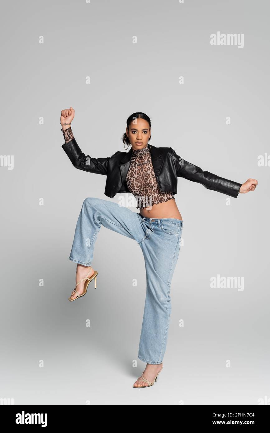 Eine lange, modische afroamerikanische Frau in hohen Absätzen und trendigen Outfits, die auf einem Bein posiert, auf grauem Stockbild Stockfoto
