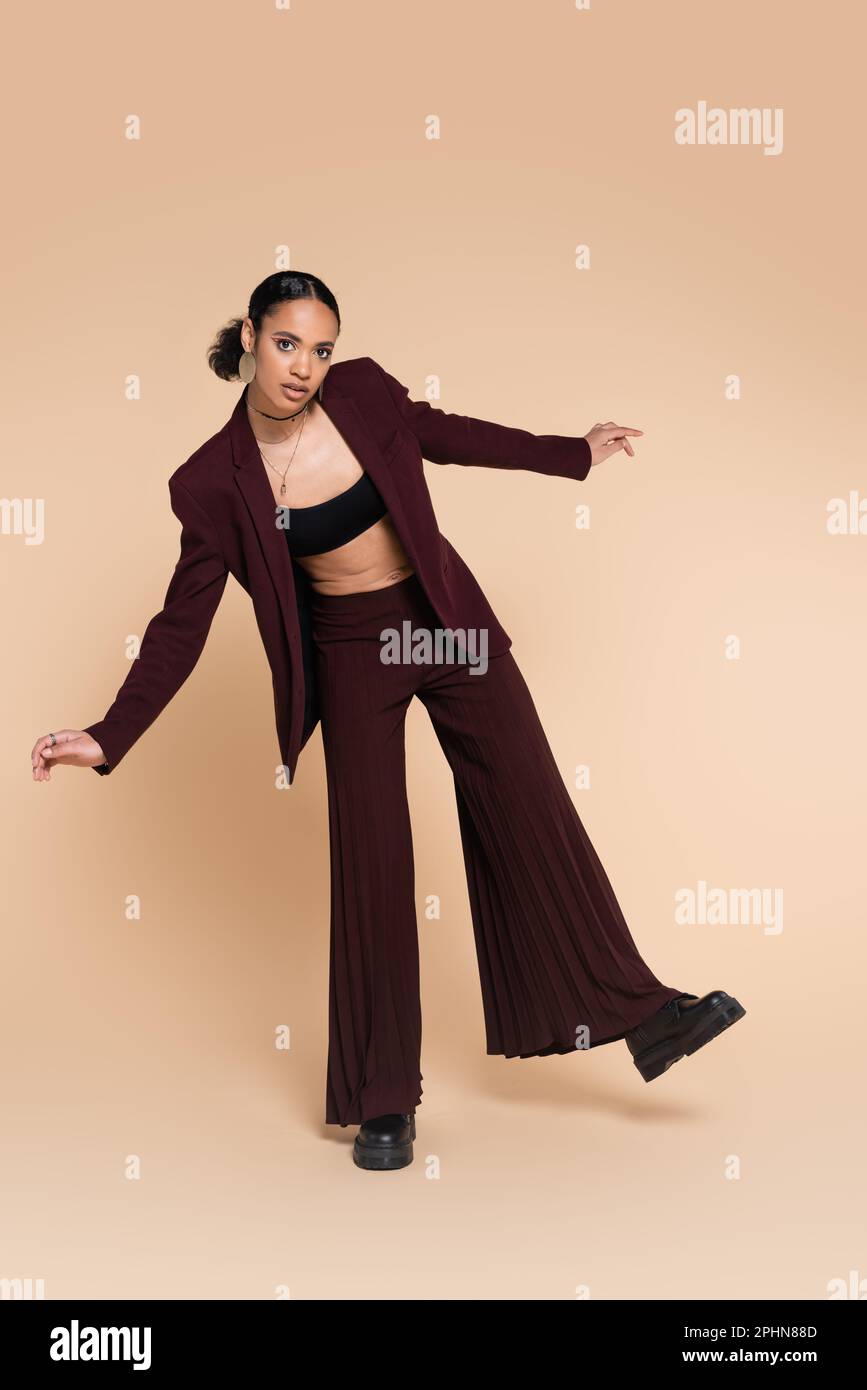 Lange, hübsche afroamerikanische Frau in kastanienbraunem Anzug mit breiter Beinhose, die auf Beige posiert, Stockbild Stockfoto
