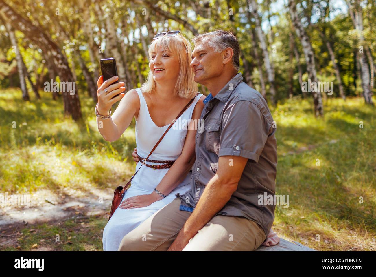 Porträt eines älteren Familienpaares, das Selfie mit dem Smartphone im Sommerwald macht. Ältere Menschen ruhen sich auf einer Bank aus und fotografieren. Mann und Frau Stockfoto