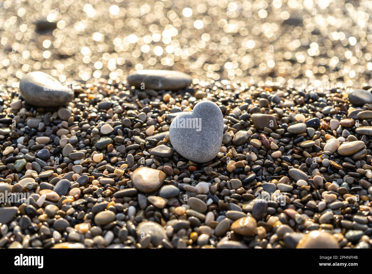 Stone Heart am Pebble Beach, Shining Ocean Water, Rocky Shore Love Symbol, Summer Pebble Heart, Urlaubskonzept, verschwommener Hintergrund Stockfoto