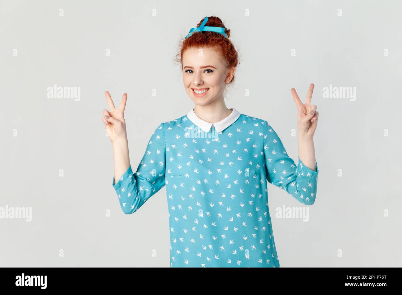 Porträt einer positiv optimistischen rothaarigen Frau mit Haarbrötchen, stehend mit V-Zeichen, mit Glück in die Kamera, blaues Kleid. Studioaufnahmen im Innenbereich isoliert auf grauem Hintergrund. Stockfoto