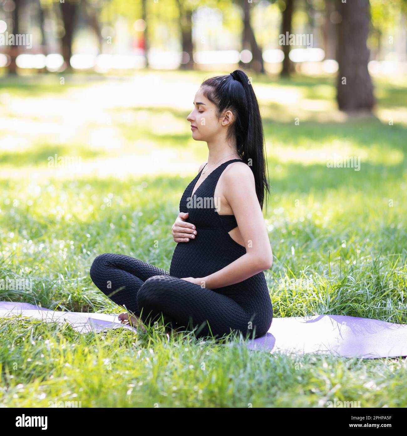 Eine junge schwangere Frau, die einen gesunden Lebensstil führt und Yoga praktiziert, meditiert in einer Lotusposition auf einer Matte im Park auf einem sonnigen warmen MOR Stockfoto