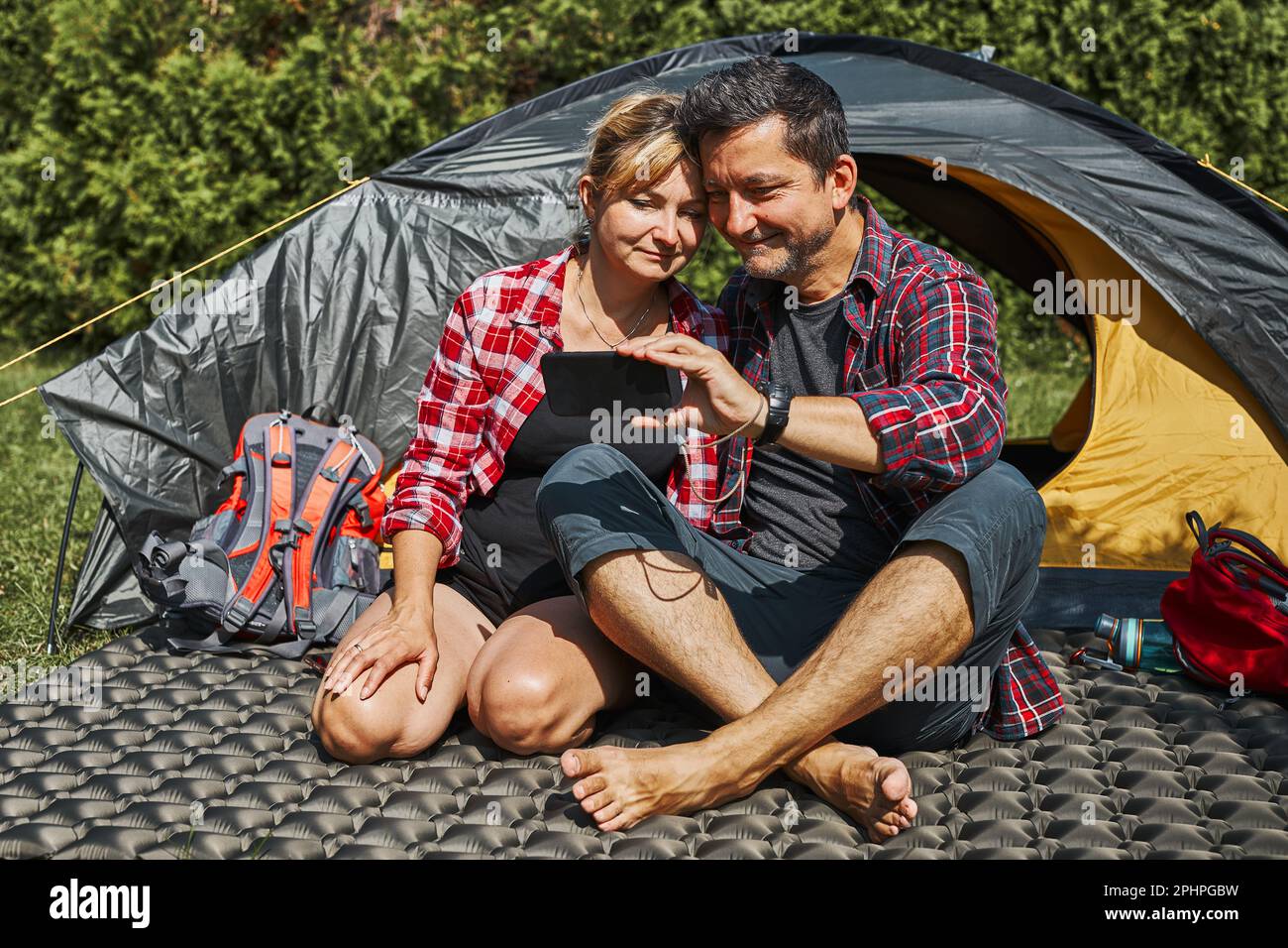 Ein Paar führt Videoanrufe mit Freunden, die ihr Smartphone verwenden, während es im Zelt beim Camping sitzt. Die Leute entspannen sich im Zelt während der Sommerferien. Aktiv spenden Stockfoto