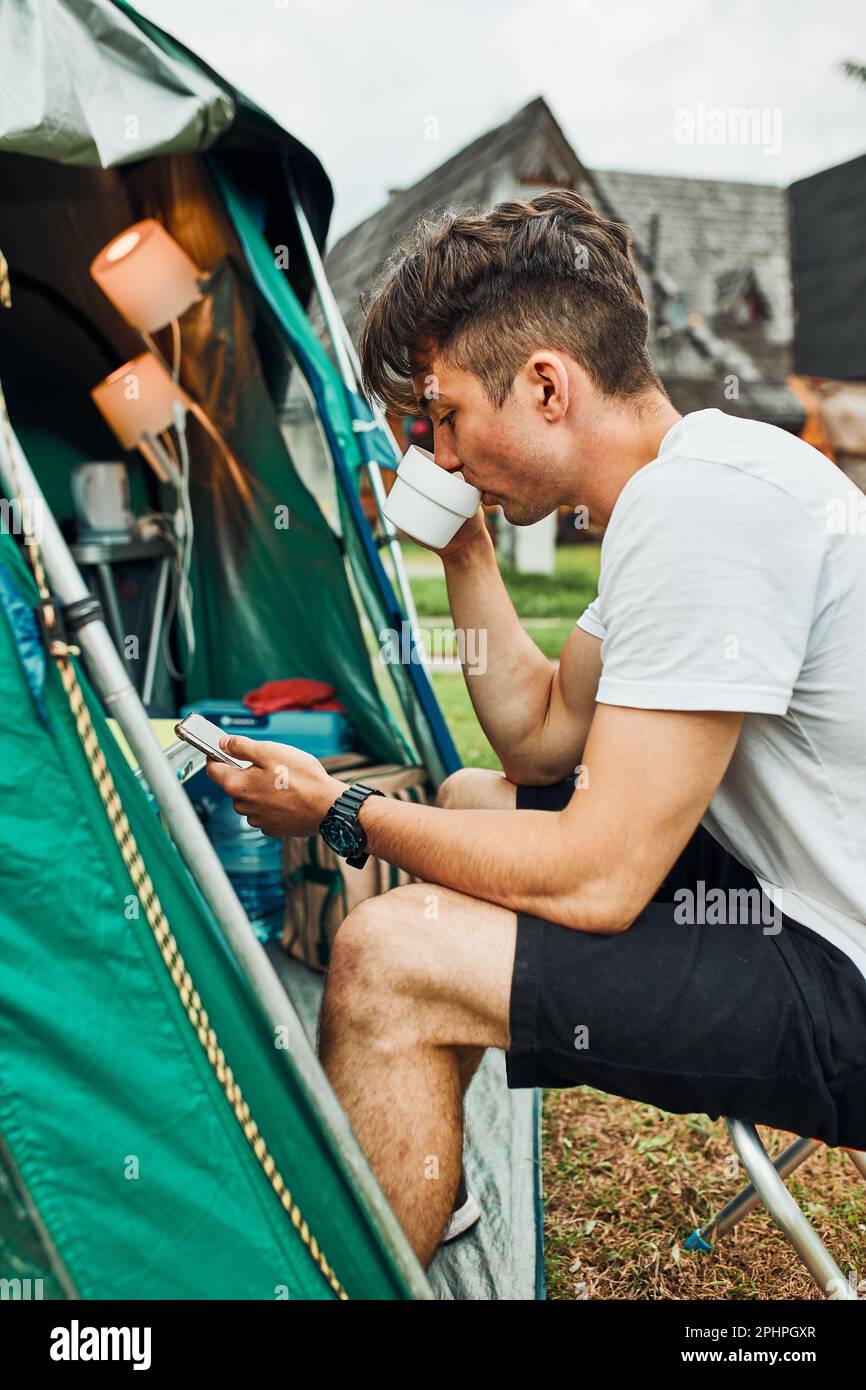 Junger Mann, der morgens einen Kaffee trinkt und vor dem Zelt sitzt. Teenager genießen Freizeit während Wochenendausflug im Sommer Stockfoto