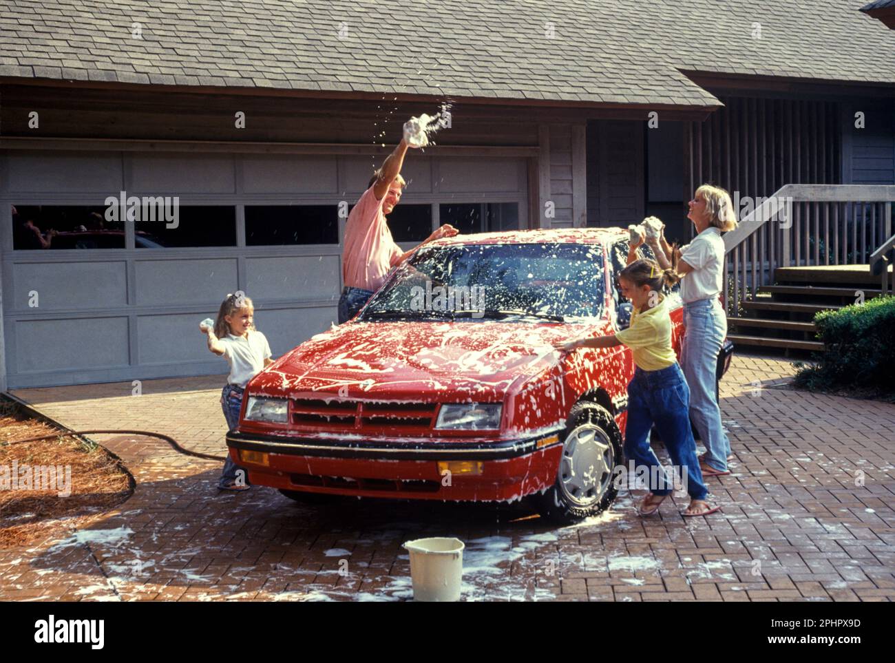 1990 HISTORISCHE FAMILIE ZUSAMMEN WASCHEN ROTES AUTO ('CHRYSLER CORP 1990) IN DER AUFFAHRT DES AMERIKANISCHEN VORSTADTHAUSES IM RANCH-STIL Stockfoto
