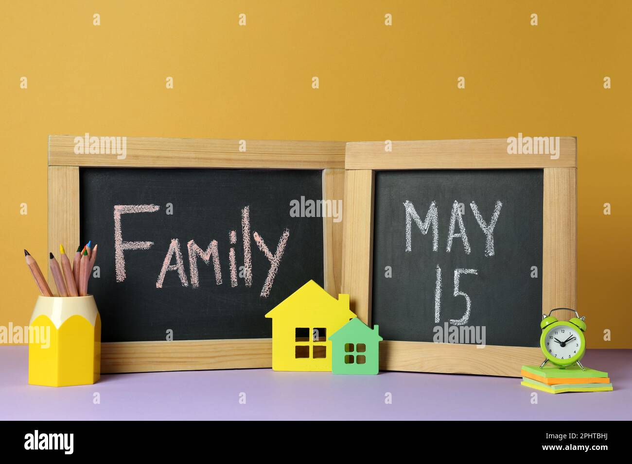 Alles Gute Zum Internationalen Familientag. Kleine Tafel mit Text, Hausmodellen, Wecker und Schreibwaren auf violettem Tisch vor gelbem Hintergrund Stockfoto