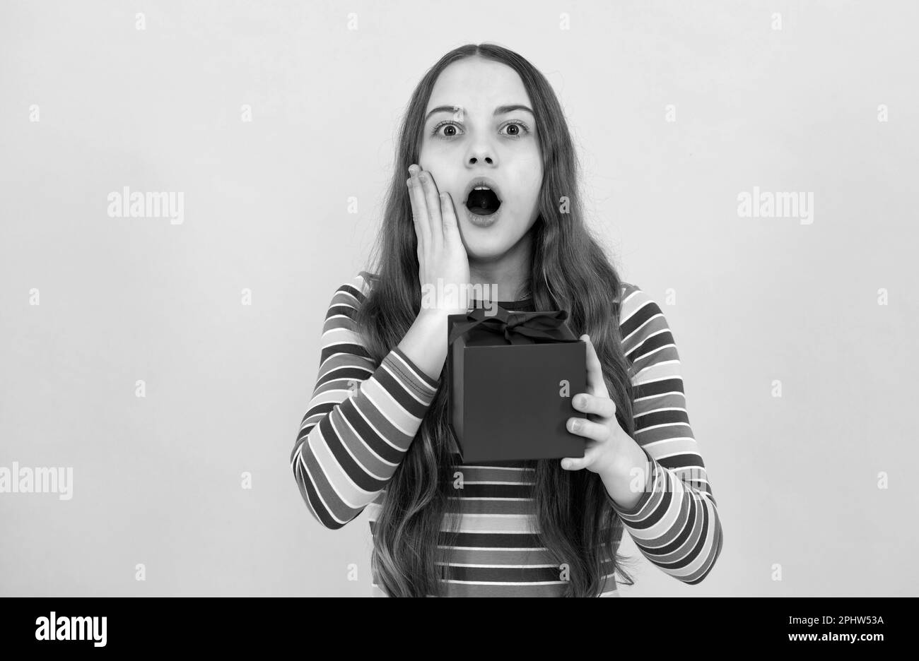 Erstaunte überraschende Emotionen eines jungen Teenagers. Ein Teenager, der eine Geschenkbox mit einem Kind auf einem gelben, isolierten Hintergrund hält. Geburtstagsgeschenk für Kinder. Weihnachten oder Stockfoto