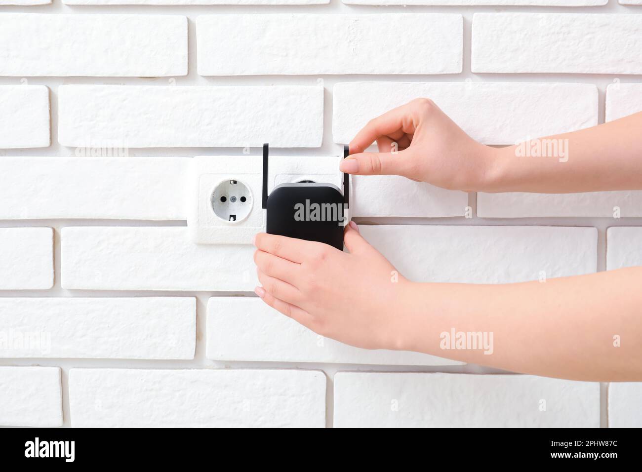 Frau, die den schwarzen WLAN-Repeater in die Steckdose an der weißen Backsteinwand steckt Stockfoto