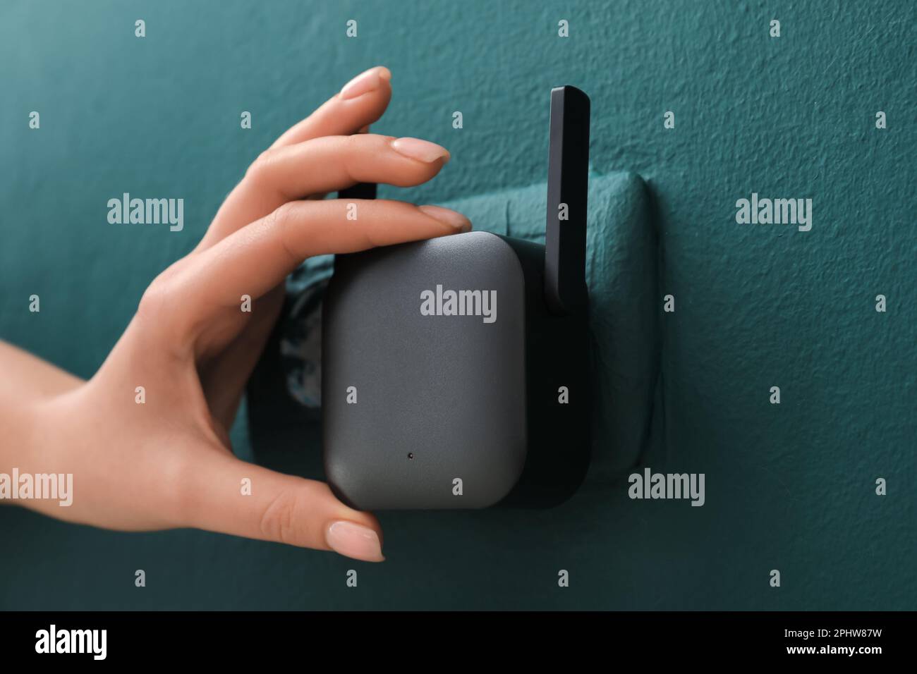 Frau, die den schwarzen WLAN-Repeater in die Steckdose an der grünen Wand steckt Stockfoto