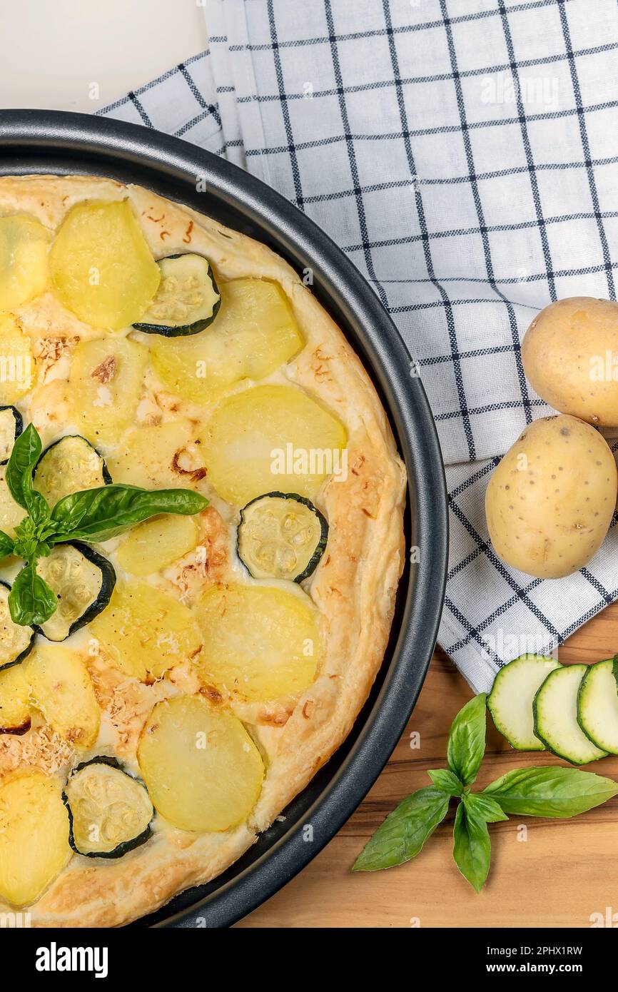 Eine Quiche mit Basilikumblättern, Kartoffeln und Zucchini in Scheiben auf einem Holzbrett mit ganzen Kartoffeln daneben Stockfoto