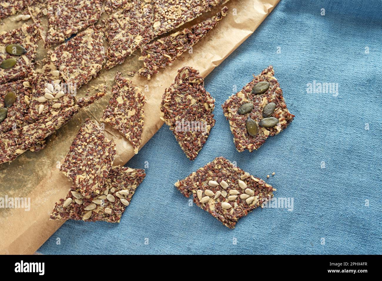 Knusprige Vollkorn-Snacks aus Hafer und Leinsamen garniert mit verschiedenen Samen auf Backpapier und einer blauen Serviette, gesundes Backen, Draufsicht von oben, Kopie Stockfoto