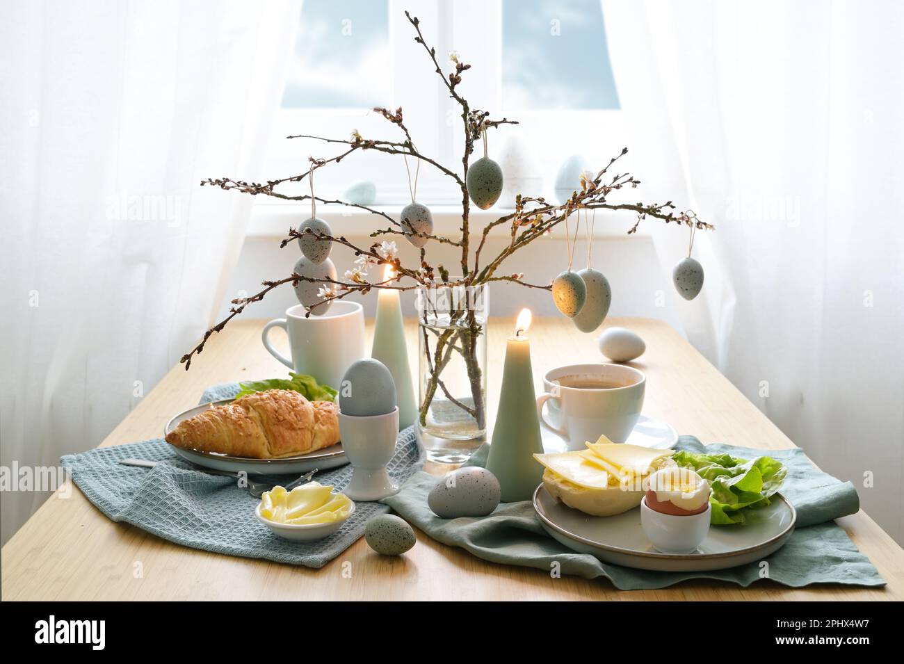 Frühstückstisch mit hängenden Ostereiern auf Federzweigen, Kerzen und blaugrauen Dekorationen vor einem Fenster mit weißen Vorhängen, Kopierbereich, s. Stockfoto