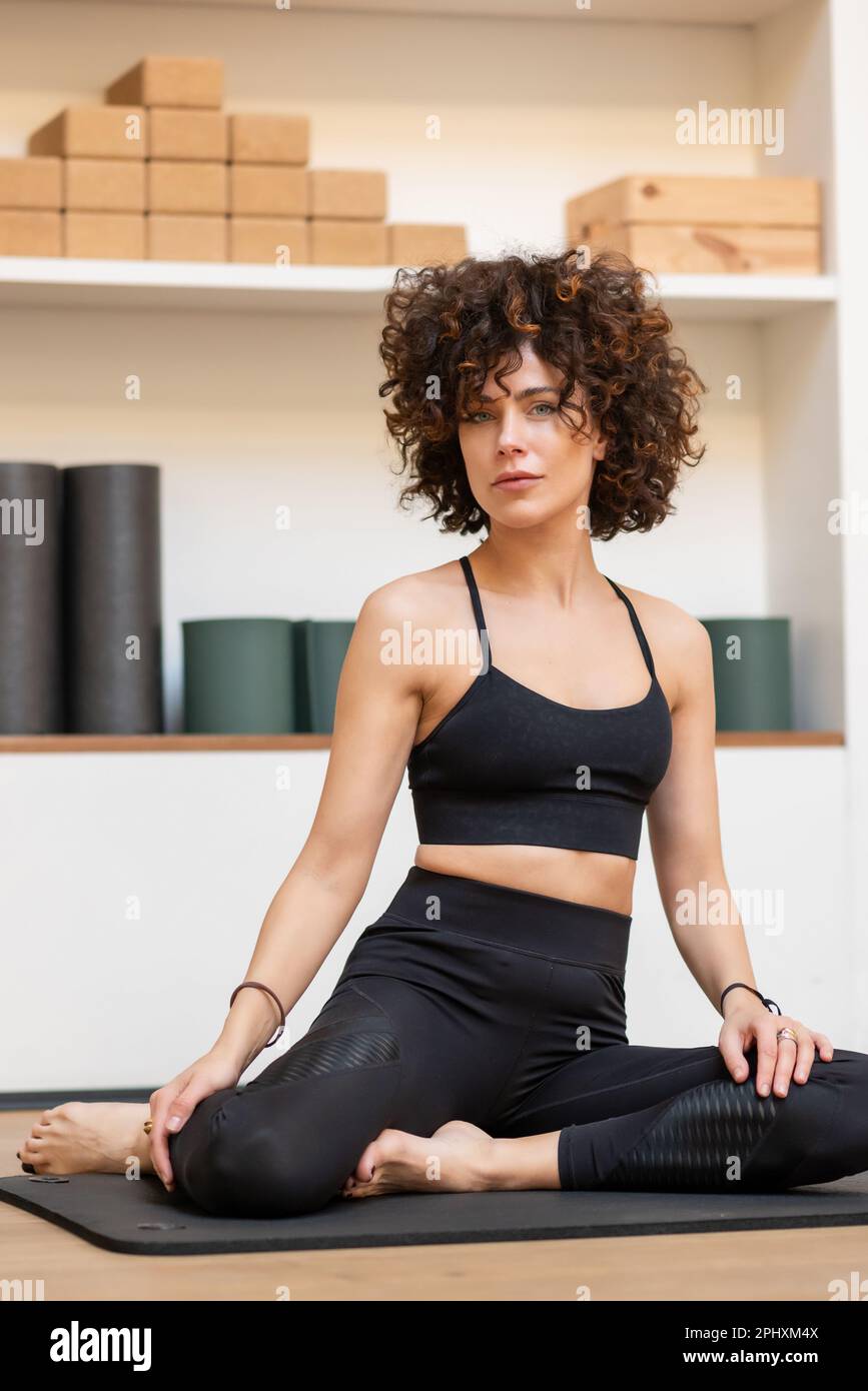 Friedlich schöne Frau in Aktivbekleidung, die auf der Matte sitzt und sich während der Yoga-Session ausruht, während sie in die Kamera schaut Stockfoto
