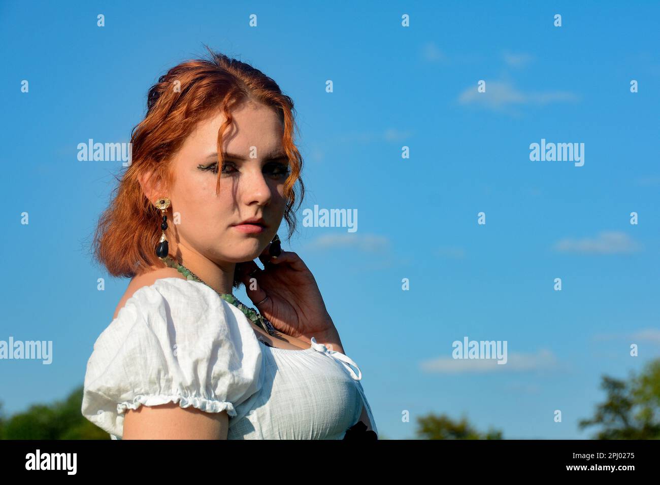 Porträt einer hübschen jungen Frau, die ein weißes Oberteil trägt, draußen mit grünen Bäumen und blauem Himmel steht Stockfoto