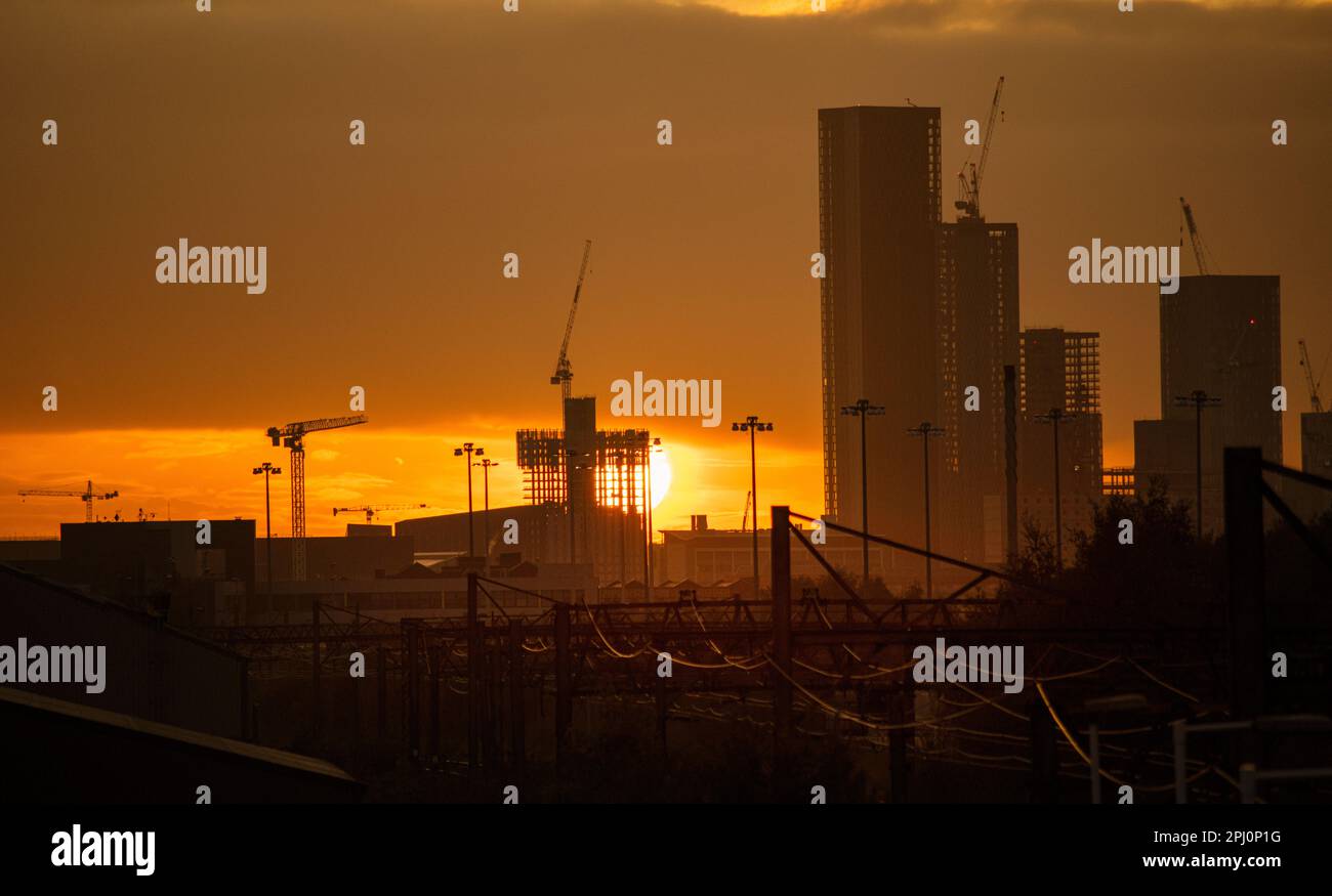 Sonnenuntergang hinter Baustellen und weit entfernt von Gebäuden der Stadt. Die Türme und Kräne von Manchester, England, mit der orangefarbenen Sonne dahinter Stockfoto