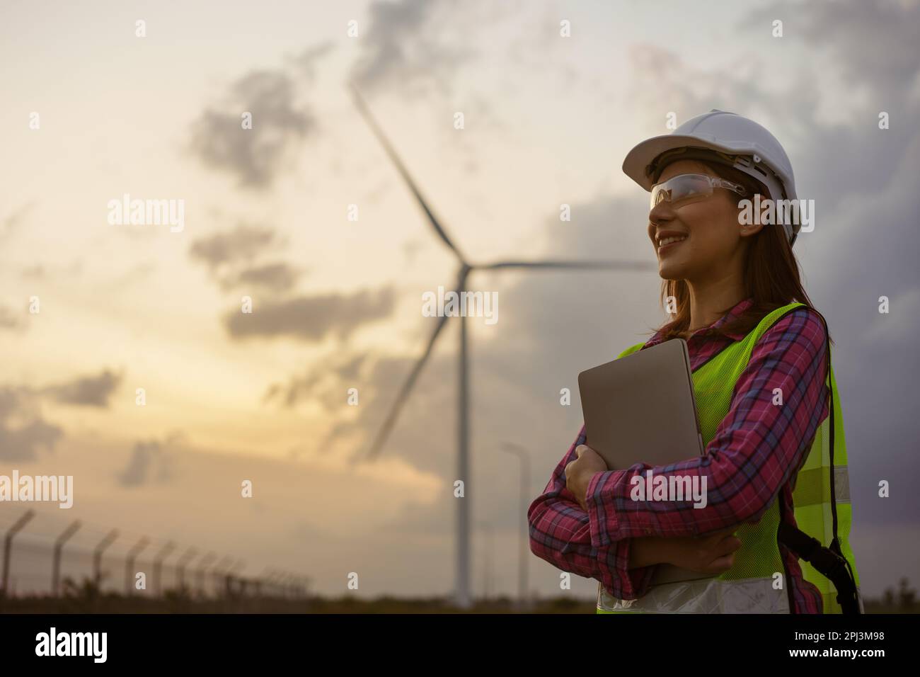 Asiatische Frau mit weißem Helm, die mit einem Laptop auf einer Farm für erneuerbare Energien arbeitet. Weibliche Inspektorin, die die Funktion von Windturbinen im Freien kontrolliert. Stockfoto